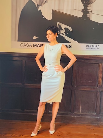 Cecilia Muilone, en vestido blanco y tacones, sonríe de pie frente a un gran póster en blanco y negro de Mariano Mores al piano. Pared y piso de madera