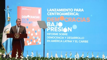 Un hombre con traje oscuro y barba gris habla desde un podio de madera frente a una pantalla azul que muestra el título "Democracias bajo presión". Una bandera de Guatemala está a su izquierda