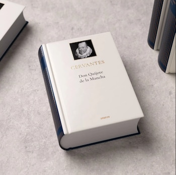 Primer plano de un libro de tapa dura blanca con el título 'Don Quijote de la Mancha' de Cervantes, con un retrato del autor y detalles dorados
