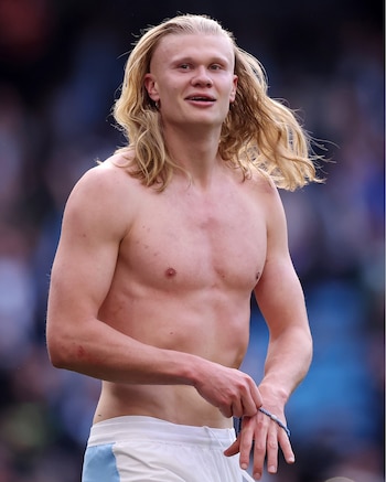 Erling Haaland, con el torso desnudo y cabello rubio largo, sonríe levemente mientras ajusta una pulsera azul en su muñeca, vistiendo shorts blancos y celestes