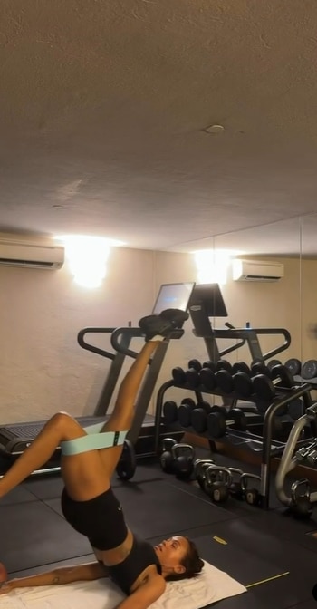Zoe Saldaña en el suelo de un gimnasio. Ella usa una banda de resistencia en sus muslos y levanta una pierna. Equipo de gimnasio visible