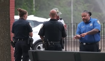Dos agentes de policía, un guardaparques y un empleado de parques conversan al aire libre. Hay un árbol, un coche blanco y una valla de fondo