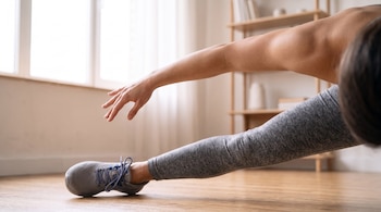 Brazo y pierna de una persona con ropa deportiva y zapatilla gris extendidos sobre un suelo de madera junto a una ventana.