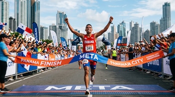 Un atleta con camiseta roja número 345 cruza la cinta de meta rodeado de público con banderas y rascacielos de fondo.