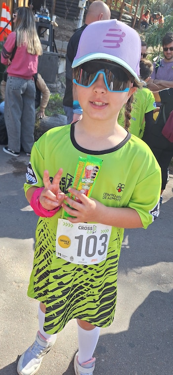 Una niña con gorra lila, anteojos de sol y camiseta deportiva verde con el número 103 sostiene un paquete de golosinas