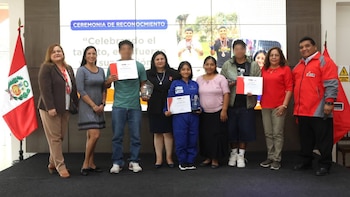Grupo de diez personas en un escenario con una bandera de Perú. Una pantalla de fondo muestra texto y fotos. Los adolescentes sostienen certificados y trofeos