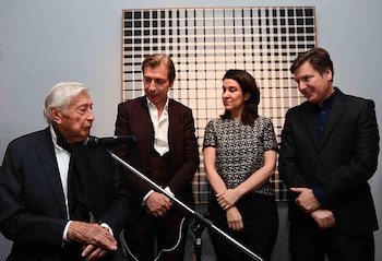 Julio Le Parc, sentado de perfil, habla por micrófono. Detrás, tres personas de pie y una obra de arte abstracto con cuadrícula en blanco y negro en la pared