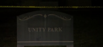 Un letrero oscuro con las palabras "UNITY PARK" en letras claras, iluminado en la oscuridad de la noche, con cinta amarilla de la policía visible en la parte superior