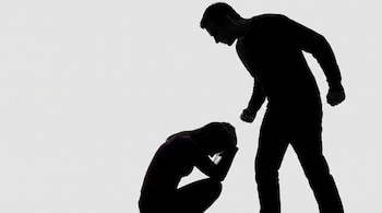 Silueta de un hombre de pie, con puño cerrado, inclinado sobre una silueta de mujer acurrucada, con las manos en la cabeza, sobre fondo blanco.