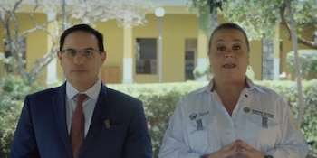 Hombre con traje azul y mujer con camisa blanca posan mirando al frente. Detrás, vegetación y un edificio amarillo con pilares blancos