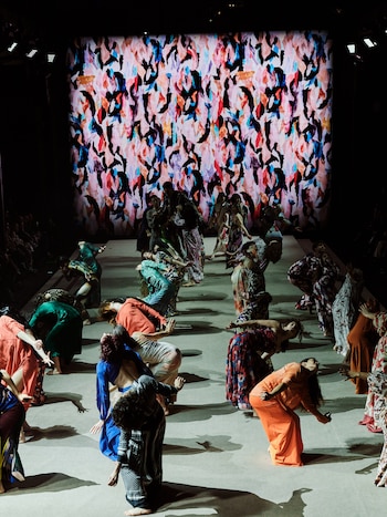 Un grupo de bailarines realiza una coreografía sobre una pasarela frente a una gran pantalla con estampado multicolor