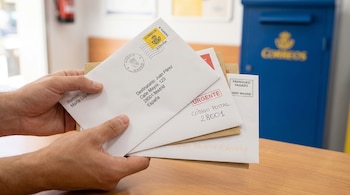 Manos sujetando varios sobres de papel con sellos de Correos de España y un código postal frente a un buzón azul.