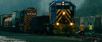 Dos trenes de carga con locomotoras azul y amarilla, y naranja y negra, en vías férreas con dos hombres caminando en primer plano