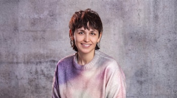 Mujer joven con cabello corto castaño, ojos marrones y aretes dorados, sonriendo, vestida con suéter de colores púrpura, rosa y blanco, sobre pared gris texturizada.