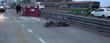 Una motocicleta roja y blanca está tendida en una carretera. Se observan un vehículo policial, dos agentes, una carpa roja y un autobús en el carril adyacente