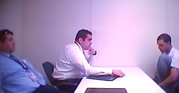 Tres hombres en una sala de paredes blancas. Dos visten camisa y están junto a una mesa, mientras el tercero permanece sentado en una silla al lado