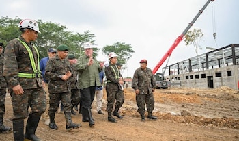 Hombres con uniformes militares y ropa civil. Cascos de seguridad. Terreno de construcción. Barro. Edificio sin terminar. Grúa roja