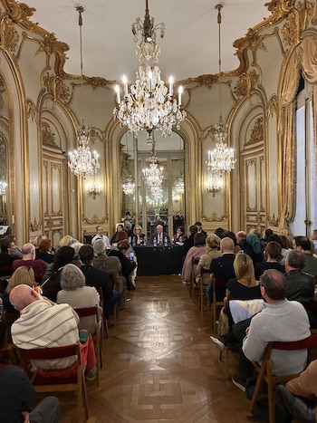 Público sentado en hileras de sillas frente a una mesa con oradores en una sala con molduras doradas y candelabros de cristal