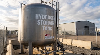 Un tanque metálico cilíndrico de almacenamiento de hidrógeno con tuberías, válvulas y manómetros, ubicado en un área industrial cercada con un edificio al fondo.