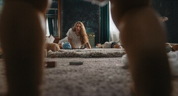 Una mujer de cabello largo en pijama está sobre una alfombra de pelo largo, rodeada de un muñeco azul, un peluche marrón y bloques. Vista baja