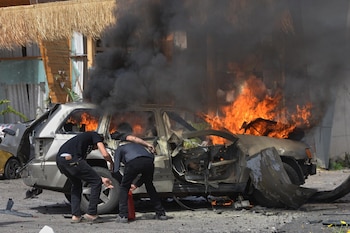 Coche gris en llamas con fuego naranja brillante y denso humo negro. Dos hombres de oscuro se agachan cerca del vehículo destruido en una calle con escombros