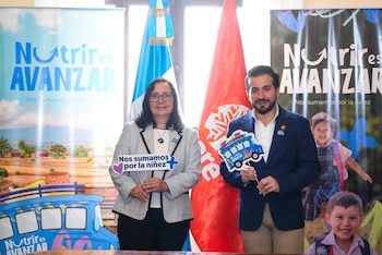 Karin Herrera y un hombre posan sonrientes en una ceremonia, cada uno sosteniendo un cartel promocional de la campaña 'Nutrir es Avanzar', con banderas y pancartas al fondo