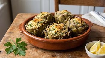 Una fuente de barro con alcauciles rellenos, un ramillete de perejil y gajos de limón en un recipiente pequeño.