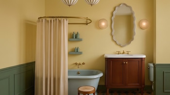Cuarto de baño con paredes amarillo y verde oscuro, bañera celeste, cortina beige, mueble de lavabo de madera, espejo de marco ondulado y luminarias.