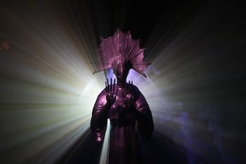 Silueta de una persona tomando una foto con un teléfono a una estatua oscura con tocado de abanico, iluminada por luz púrpura en un espacio oscuro