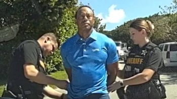 Imagen de video mostrando a Tiger Woods, vestido con un polo azul, siendo esposado por dos agentes de policía, un hombre y una mujer, al aire libre