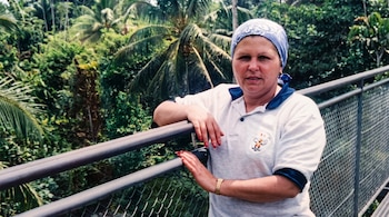 Una mujer con pañuelo azul y camiseta polo se apoya en una barandilla metálica frente a un fondo de vegetación y palmeras.