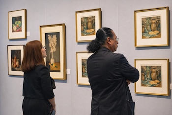 Una mujer y un hombre de espaldas observan pinturas enmarcadas colgadas en una pared gris dentro de una sala de exposición.