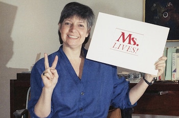 Una mujer con camisa azul sonríe, sostiene un cartel blanco con la leyenda Ms. LIVES! y realiza la señal de la victoria