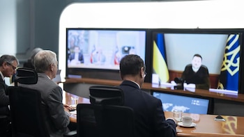Hombres de espaldas en sala de reuniones observan dos pantallas: una con el presidente Zelensky y banderas ucranianas, otra con un grupo y banderas chilenas