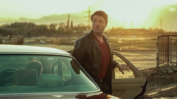 Tom Burke, un hombre de mediana edad con chaqueta de cuero y camisa roja, saliendo de un coche vintage marrón bajo un cielo dorado de atardecer