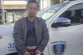Hombre con rostro difuminado, esposado, de pie frente a un vehículo policial blanco que muestra el texto "Fuerza Pública" y un escudo.