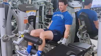 Mark Wahlberg, con camiseta azul y shorts oscuros, realiza un ejercicio de piernas en una máquina de gimnasio gris, mostrando concentración