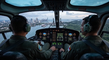 Vista de la cabina de un helicóptero con dos pilotos uniformados de espaldas frente al panel de instrumentos y la Ciudad de Panamá.