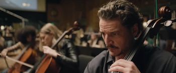Pedro Pascal toca un violonchelo en primer plano, con otras personas y violonchelos al fondo en una sala oscura