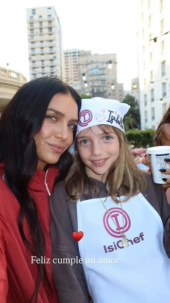 Zaira Nara posa al aire libre junto a una niña que viste un delantal y un pañuelo de cocina blancos, frente a edificios urbanos