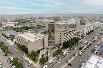 Vista aérea de un complejo de edificios grandes, calles con vehículos, autobuses y camiones, y estacionamientos llenos de automóviles bajo un cielo nublado