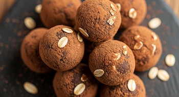 Primer plano de varias trufas redondas de cacao y avena caseras, cubiertas con cacao en polvo y adornadas con hojuelas de avena, sobre una tabla oscura.