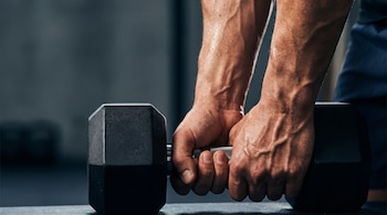 Dos manos con sudor sujetan una mancuerna negra en una superficie plana dentro de un gimnasio.