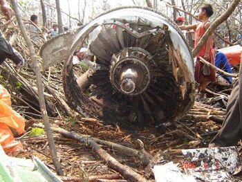 Un motor de avión destruido y partes del fuselaje yacen entre ramas de árboles junto a personas en un área de vegetación