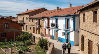 Vista aérea de un pueblo con casas de piedra, tejados rojos, calles, dos mujeres mayores caminando, bicicletas aparcadas, ropa tendida y un huerto urbano bajo cielo azul.