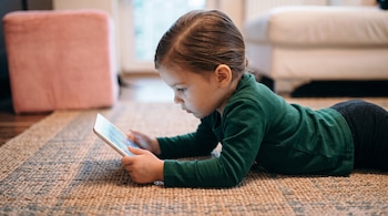 Niña vestida con camiseta verde acostada sobre una alfombra tejida mientras sostiene una tableta digital con ambas manos.