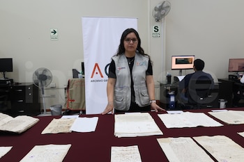 Una mujer con chaleco del AGN, documentos antiguos sobre una mesa, letreros de seguridad, ventiladores y monitores de computadora