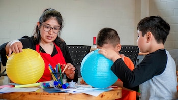 Una mujer con gafas y mandil rojo ayuda a dos niños a elaborar faroles de papel amarillo y azul en una mesa con útiles escolares