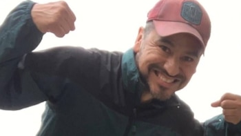 Primer plano de un hombre caucásico sonriendo a la cámara y flexionando los brazos, con una gorra roja y una chaqueta verde oscuro