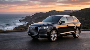 Un SUV Audi Q7 negro estacionado en una carretera asfaltada junto a una costa rocosa. Un faro se ve a lo lejos bajo un cielo de atardecer con tonos naranja y rosa.